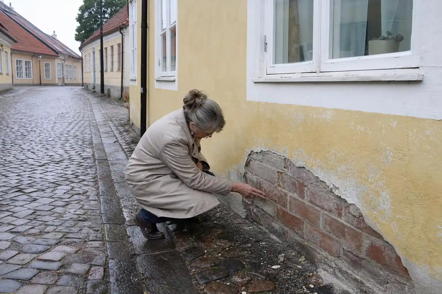 Kvinna på huk på en kullerstensgata känner på teglet där den gula putsen har släppt vid sockeln på ett lågt putsat hus
