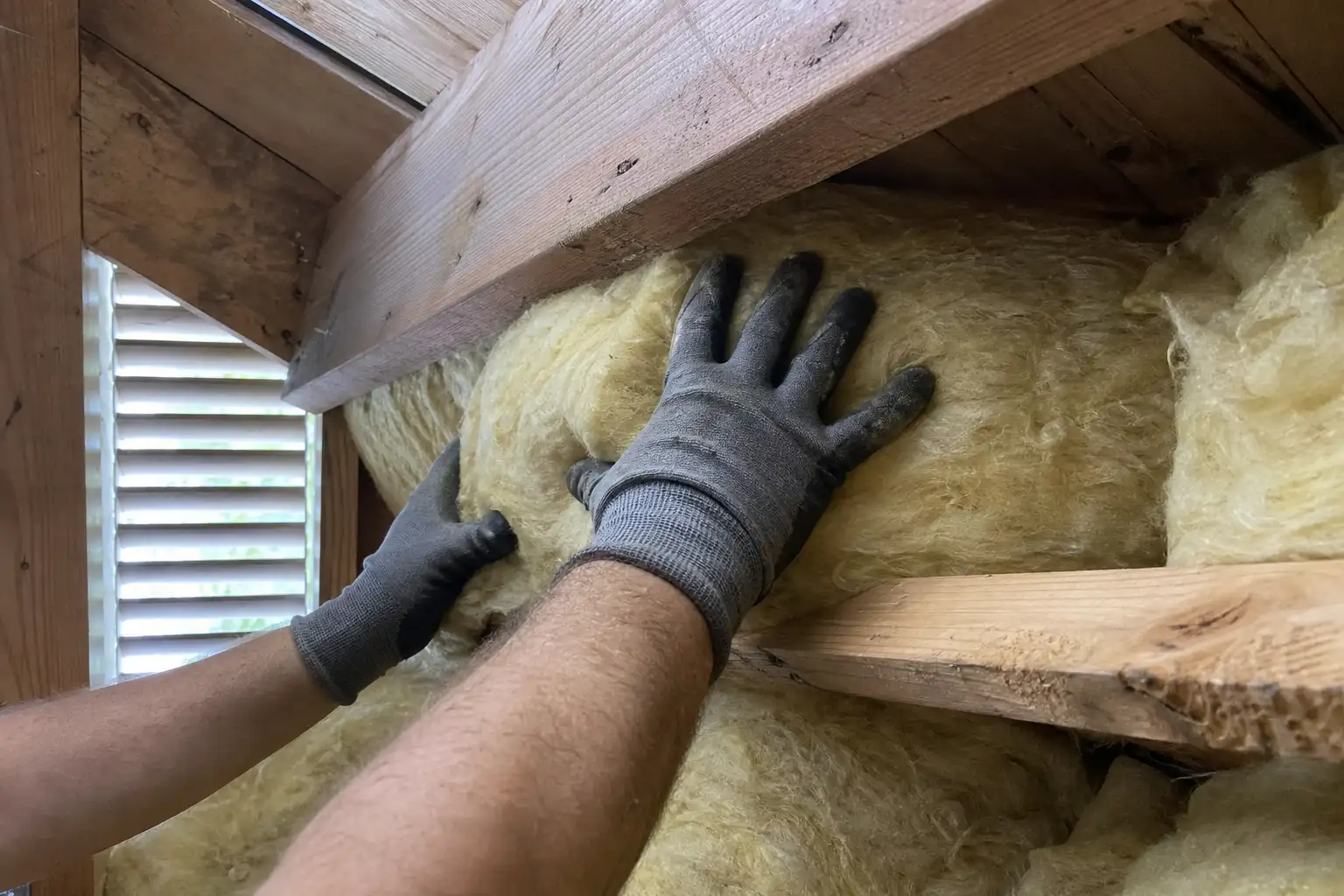 Handskbeklädda händer trycker in mineralull mellan takbjälkar, ett vardagligt exempel på arbete med klimatkrav byggnader.